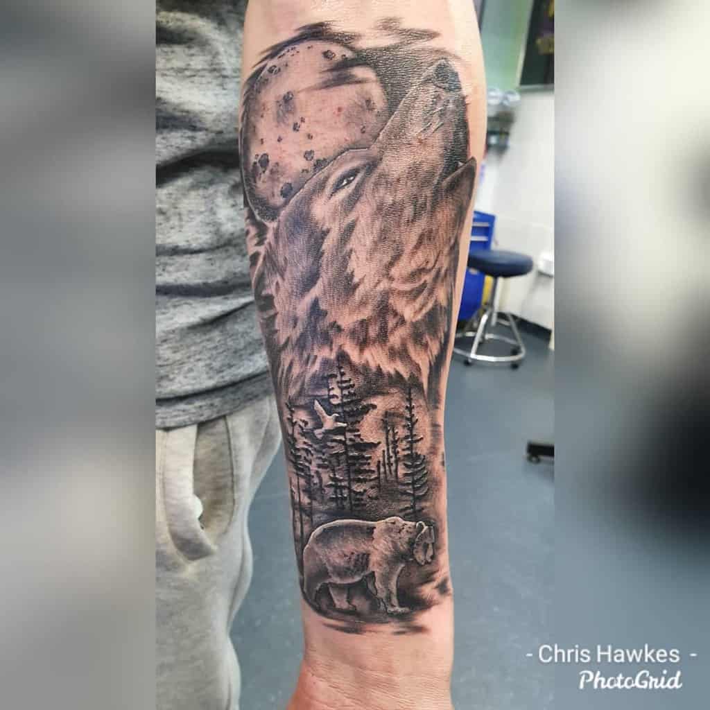 Black and grey forearm tattoo featuring a howling wolf, a full moon, trees, and a bear, creating a wilderness scene