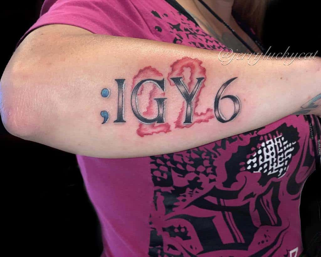 Forearm tattoo featuring ';IGY6' with a blue semicolon and red 6, designed with a blood-like effect symbolizing support and resilience