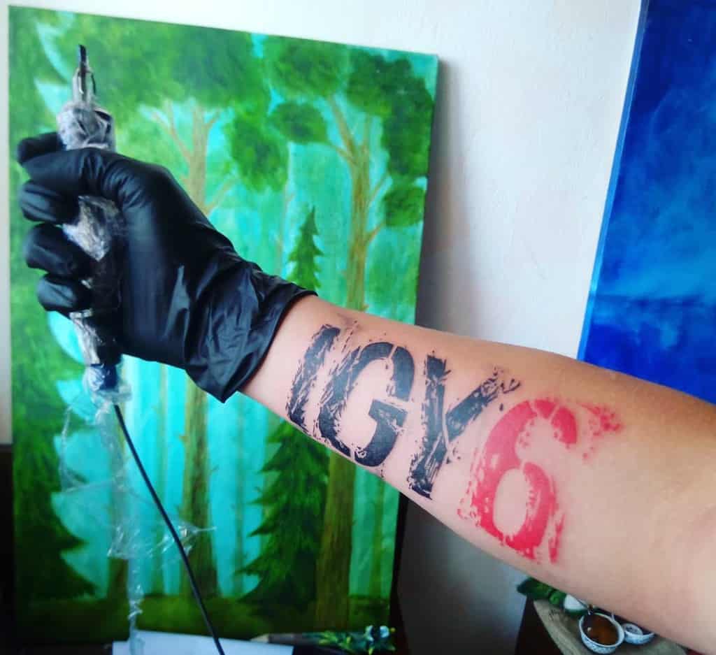 A gloved hand holding a tattoo machine next to a forearm tattoo reading "IGY6"