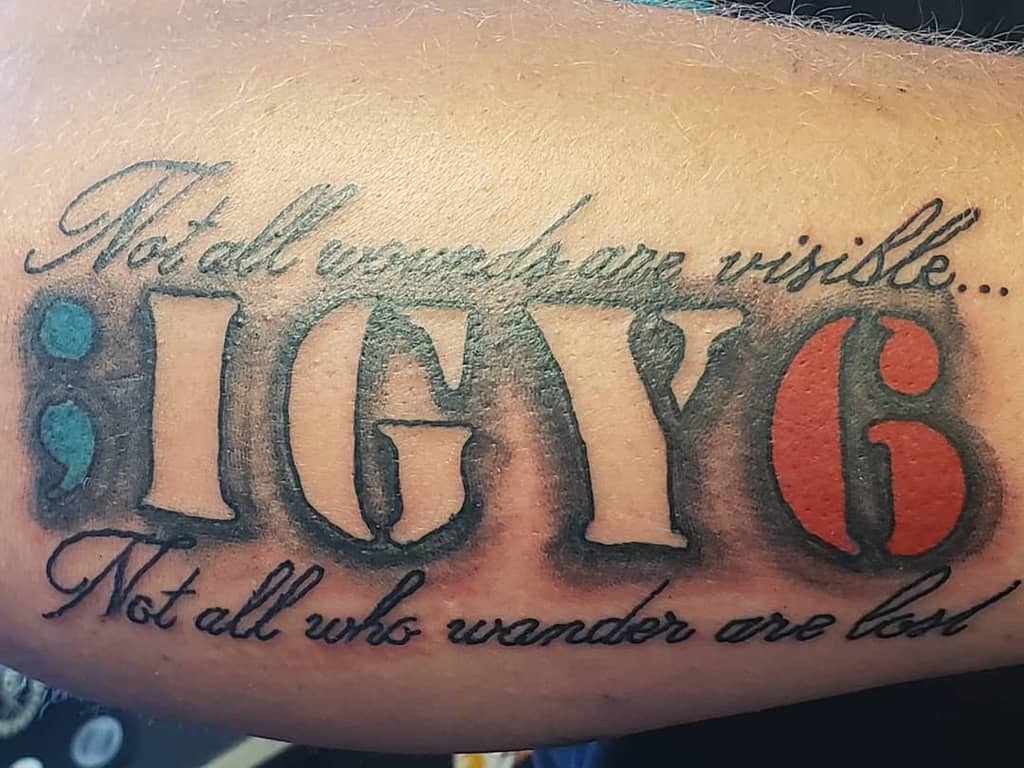 Forearm tattoo featuring ';IGY6' with a blue semicolon, red 6, and phrases 'Not all wounds are visible...' and 'Not all who wander are lost'