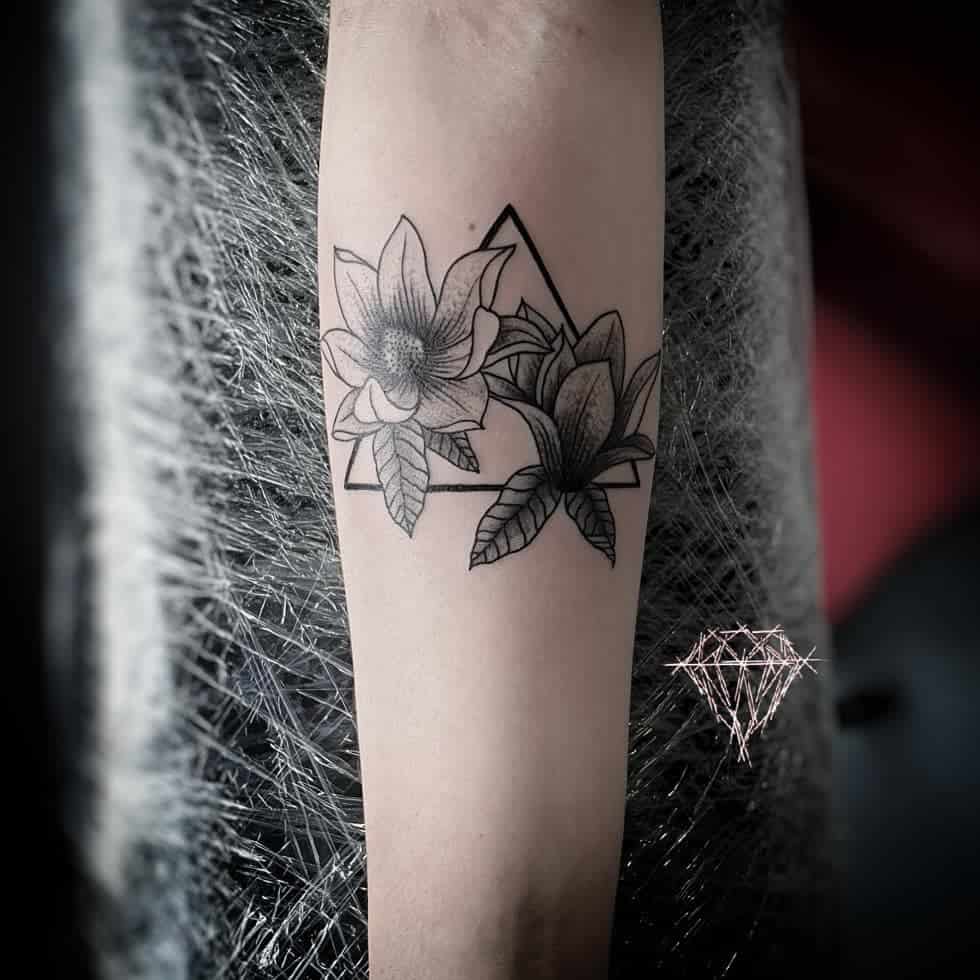 Arm tattoo featuring magnolia blossoms elegantly intertwined with a geometric triangle design, accented by a diamond-like shape nearby