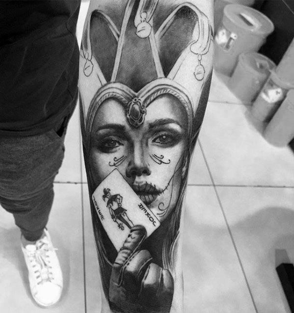A black and gray tattoo of a woman with a jester crown holding a playing card, displaying intricate facial details and expressive features.