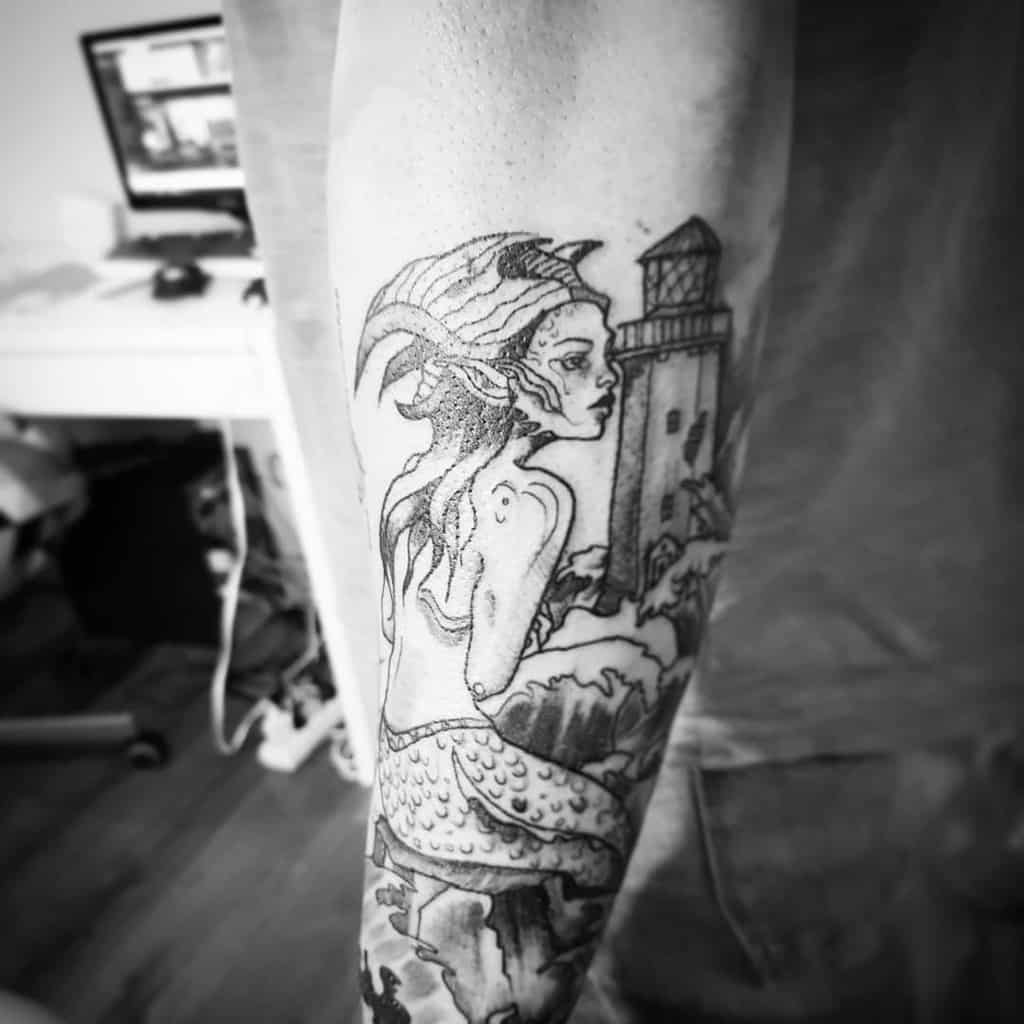 Black and grey tattoo of a mystical mermaid with horn-like fins, sitting on rocks near a lighthouse, gazing into the distance.