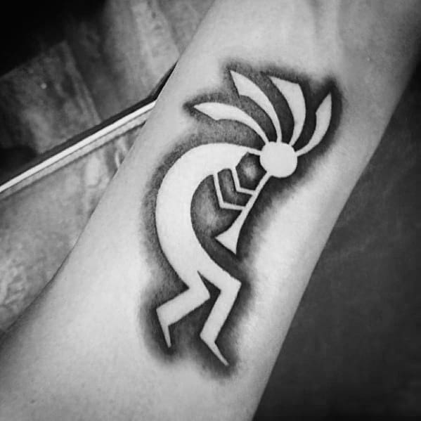 Black and white Kokopelli tattoo with bold negative space and soft shaded background, creating a striking silhouette effect on the arm.