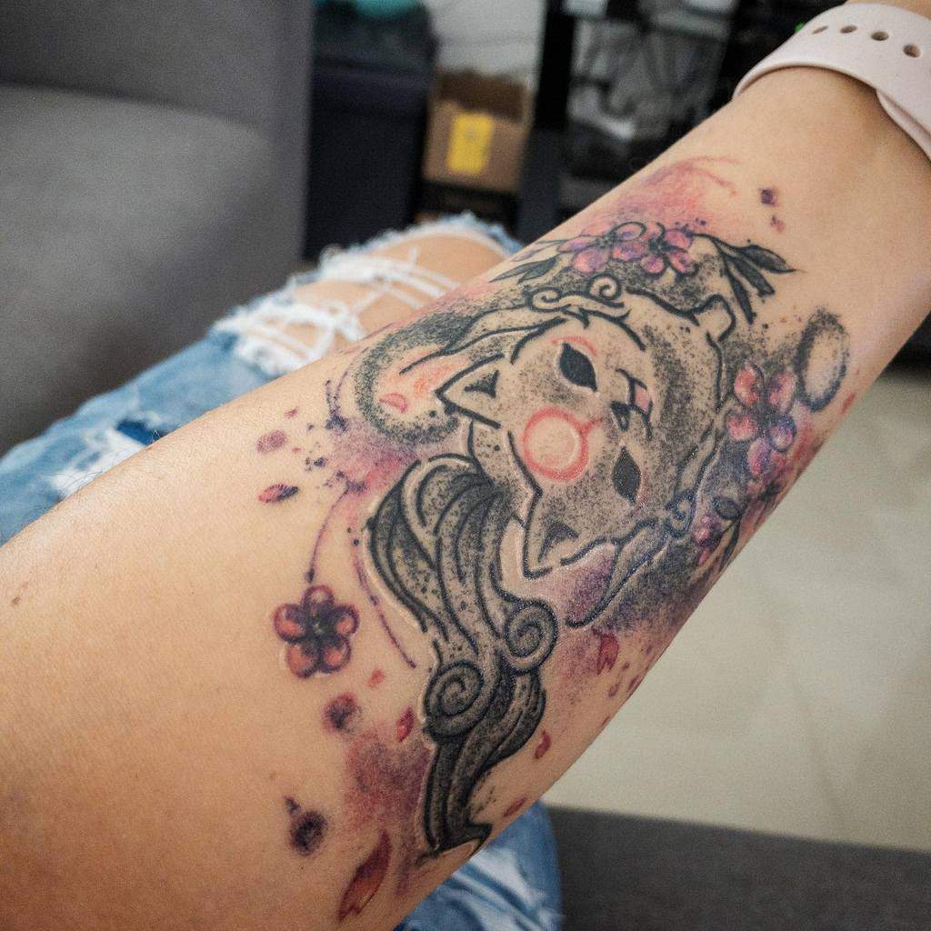 Artistic Okami tattoo on the forearm featuring Amaterasu with soft watercolor shading, cherry blossoms, and a mystical ink-wash effect