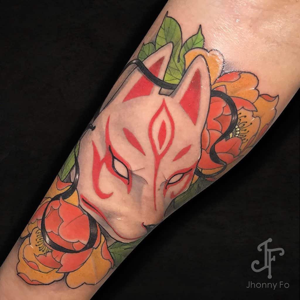 Bold Okami tattoo on the forearm featuring a Kitsune mask with striking red markings, vibrant orange flowers, and intricate black ribbon details