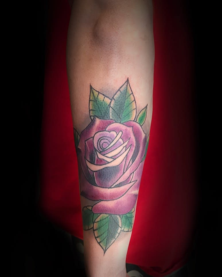 A tattoo featuring a striking purple rose with lush green leaves adorns a forearm