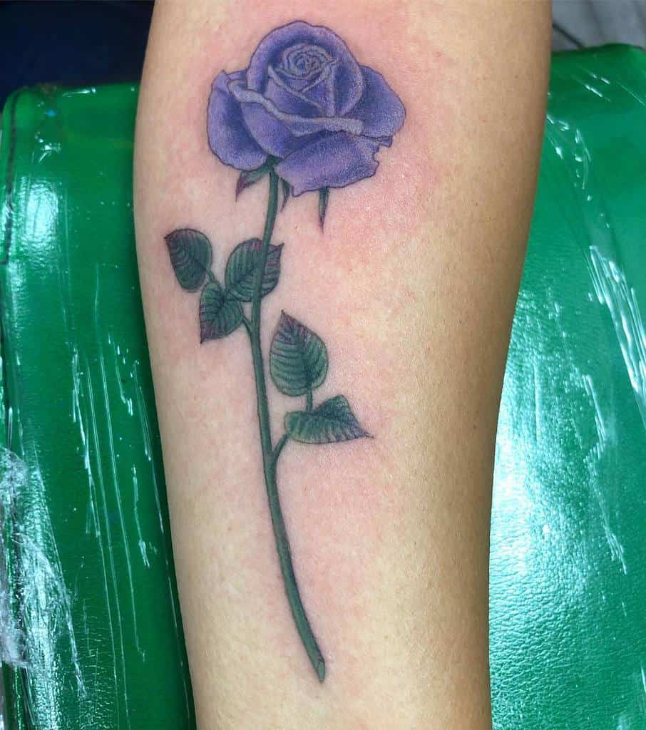 A tattoo featuring a captivating purple rose with a long, elegant stem and lush leaves graces the arm