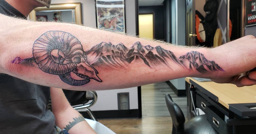 Forearm tattoo of a ram skull intricately designed with mountain range scenery