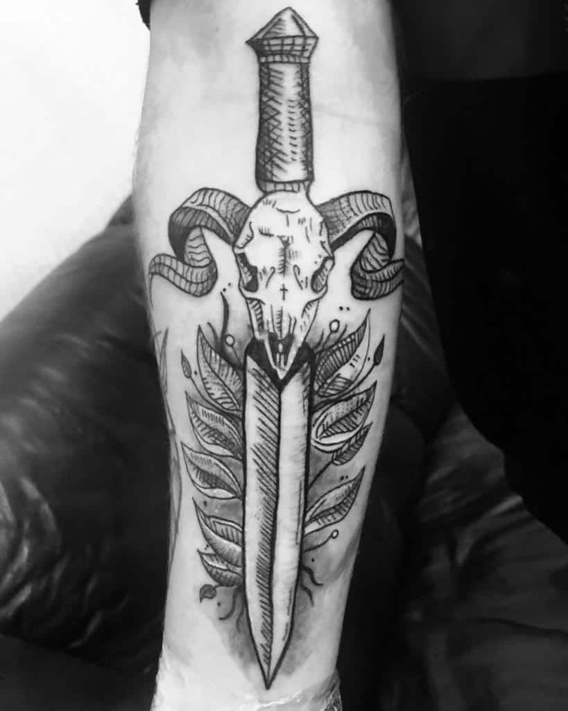 Black and white tattoo of a dagger entwined with leaves and a detailed ram skull on a forearm