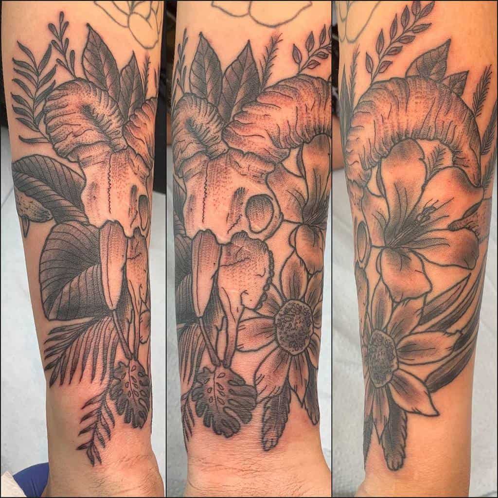 Forearm tattoo of detailed flowers and leaves in black and gray shading, accented with a striking ram skull