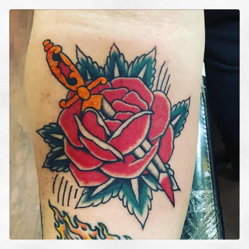 Tattoo of a dagger piercing a red rose with green leaves on skin