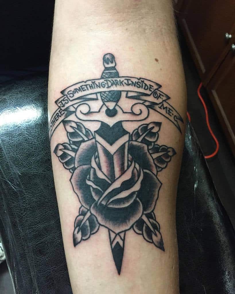 Tattoo of a dagger through a rose with a banner reading "There is something dark inside of me" on a forearm