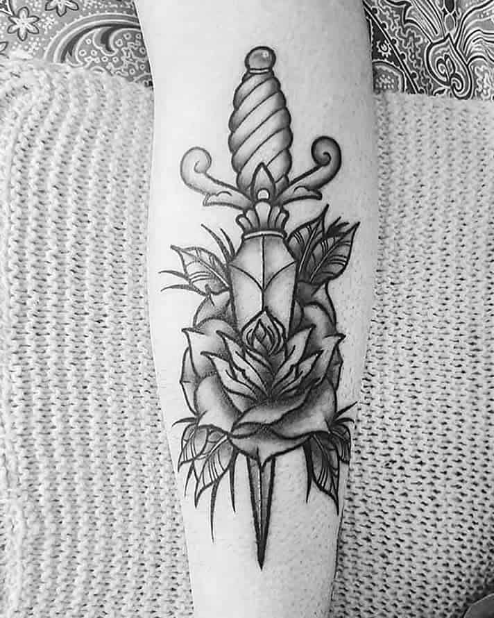 Tattoo of a dagger piercing a rose with leaves on the arm