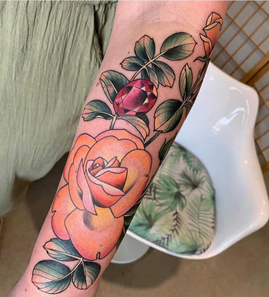 Vibrant forearm tattoo featuring a bold orange rose with lush green leaves and a detailed red gemstone accent for added charm
