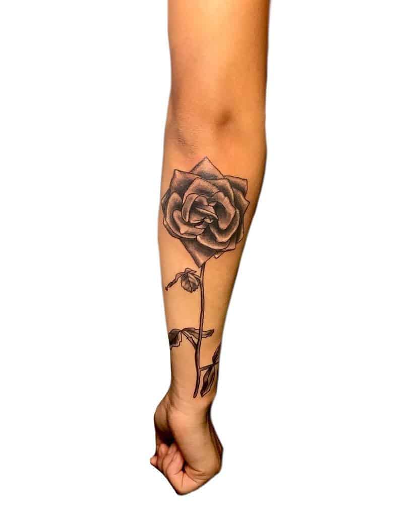 Forearm tattoo of a detailed black and gray rose with leaves and a graceful stem extending elegantly along the person's arm