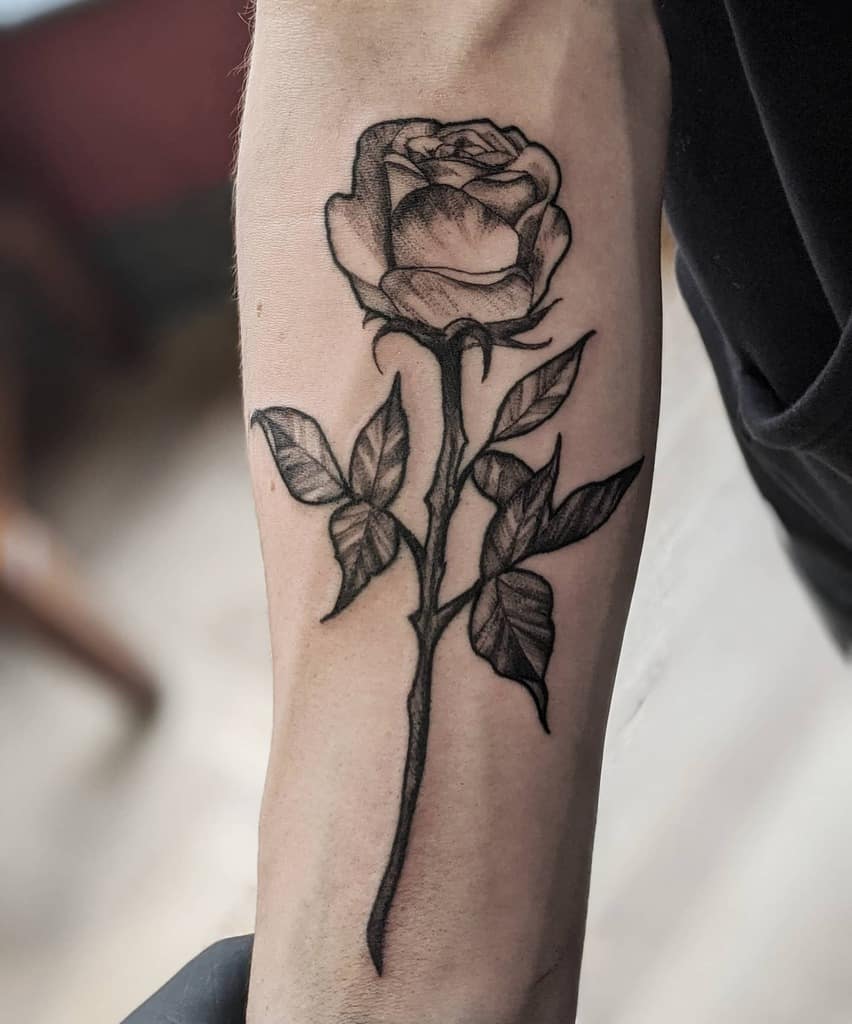 A black and gray rose tattoo elegantly winds along a person's forearm, its intricate petals flowing seamlessly into an artfully shaded stem