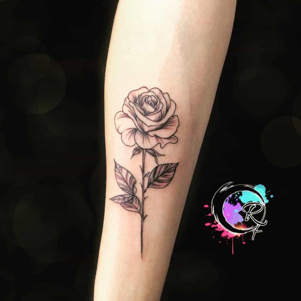 The elegant black and white rose tattoo gracefully spans the forearm, its intricate stem weaving artfully along the skin; nearby, vibrant colored initials add a striking contrast to this monochrome masterpiece