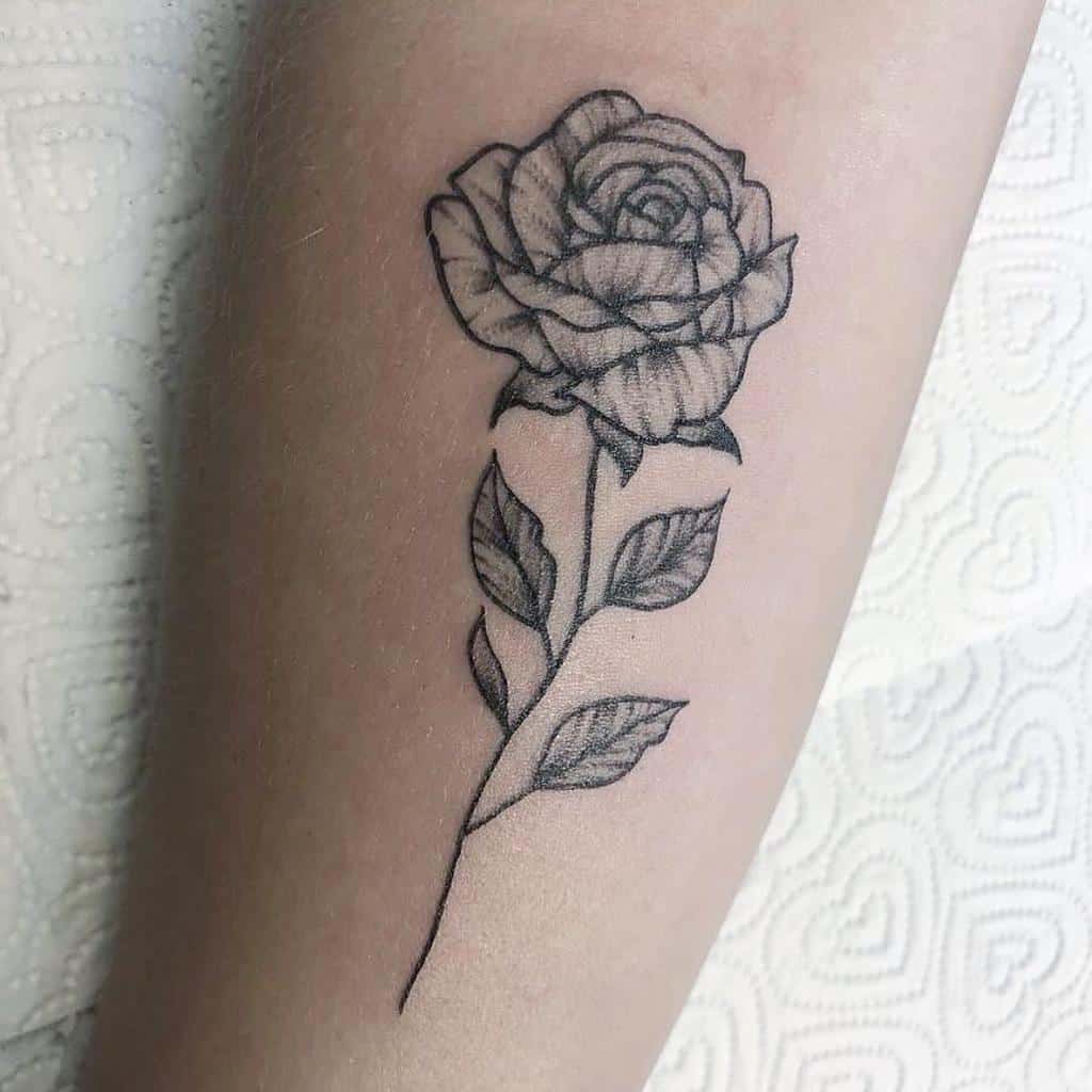 Detailed black and gray tattoo of a single rose with a long stem and leaves, placed elegantly on the forearm