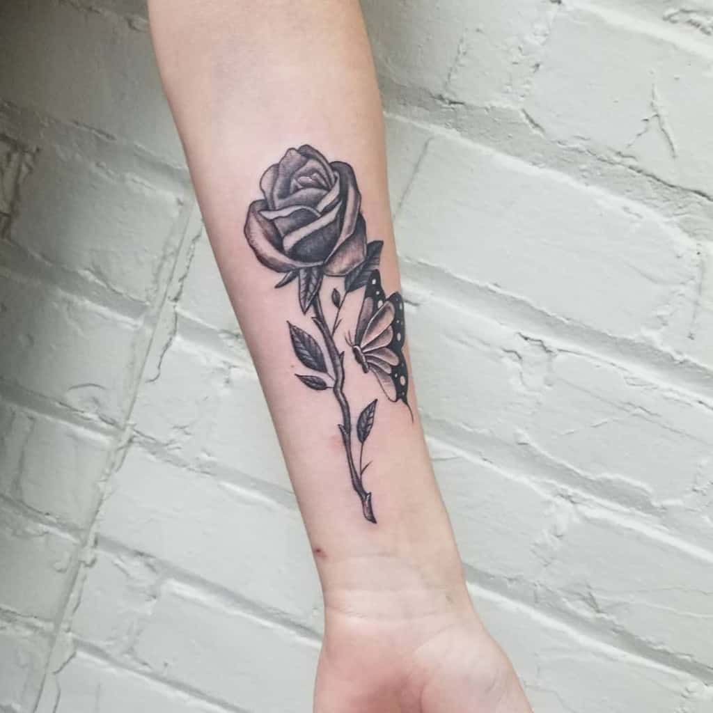 A striking black rose with an intricate stem entwines with a delicate butterfly tattoo on a person's forearm
