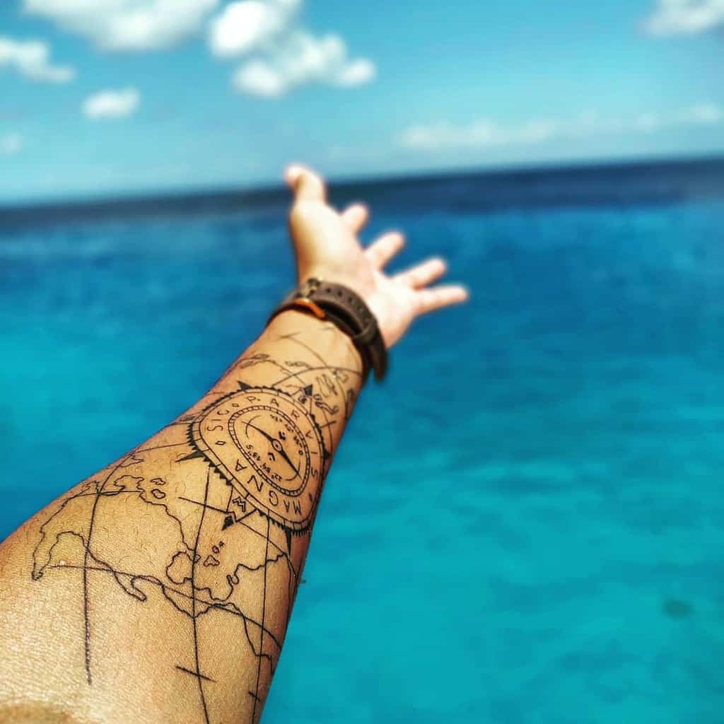 Forearm tattoo with a detailed map and compass featuring 'Sic Parvis Magna,' captured against a vibrant ocean backdrop
