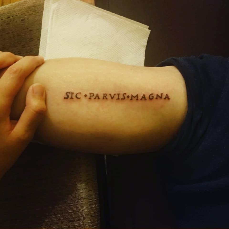 Forearm tattoo reads "Sic Parvis Magna" in Roman-style font with a hand resting on the arm