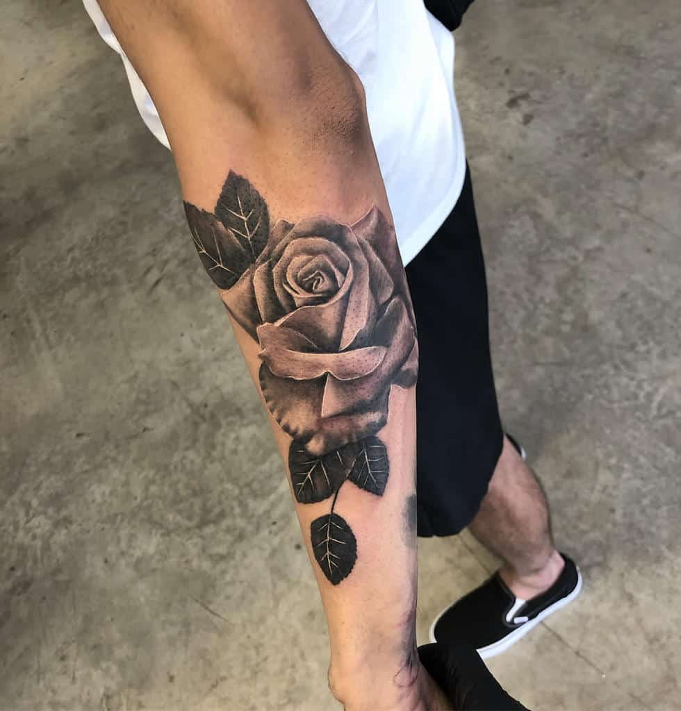 Arm tattoo of a detailed rose with leaves in black and gray ink