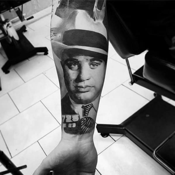 Al Capone tattoo featuring a black-and-white portrait of the gangster wearing his signature fedora, with a close-up of a stack of cash beneath him.