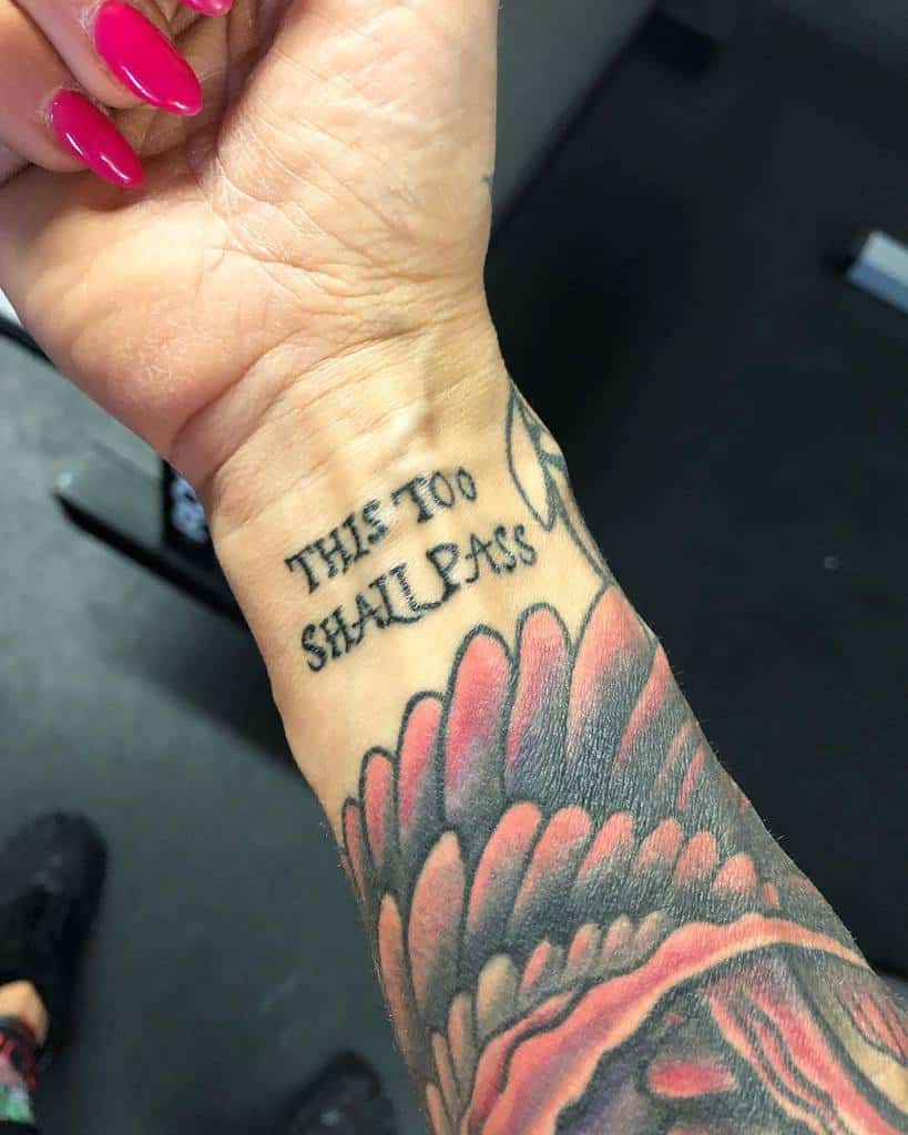 A "This Too Shall Pass" tattoo on a person's wrist, with vibrant floral tattoos surrounding it. The person has pink manicured nails.