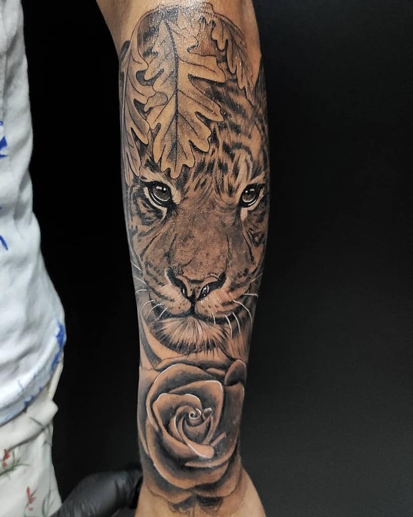 Forearm tattoo showcasing a tiger's fierce gaze intertwined with a delicate rose and majestic oak leaf, all set against a moody background
