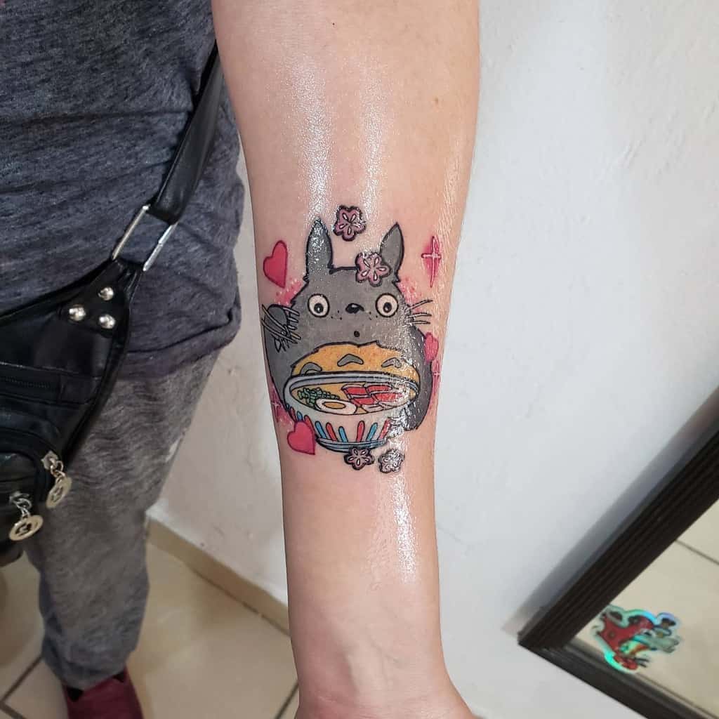 Colorful Totoro tattoo on the forearm, featuring Totoro holding a bowl of ramen with hearts, flowers, and sparkles surrounding him