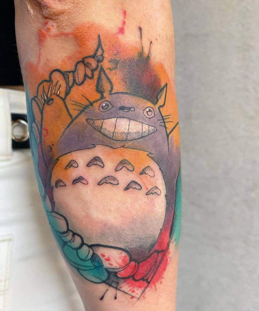 Abstract-style Totoro tattoo on the forearm, featuring Totoro with vibrant watercolor splashes in red, orange, and teal hues