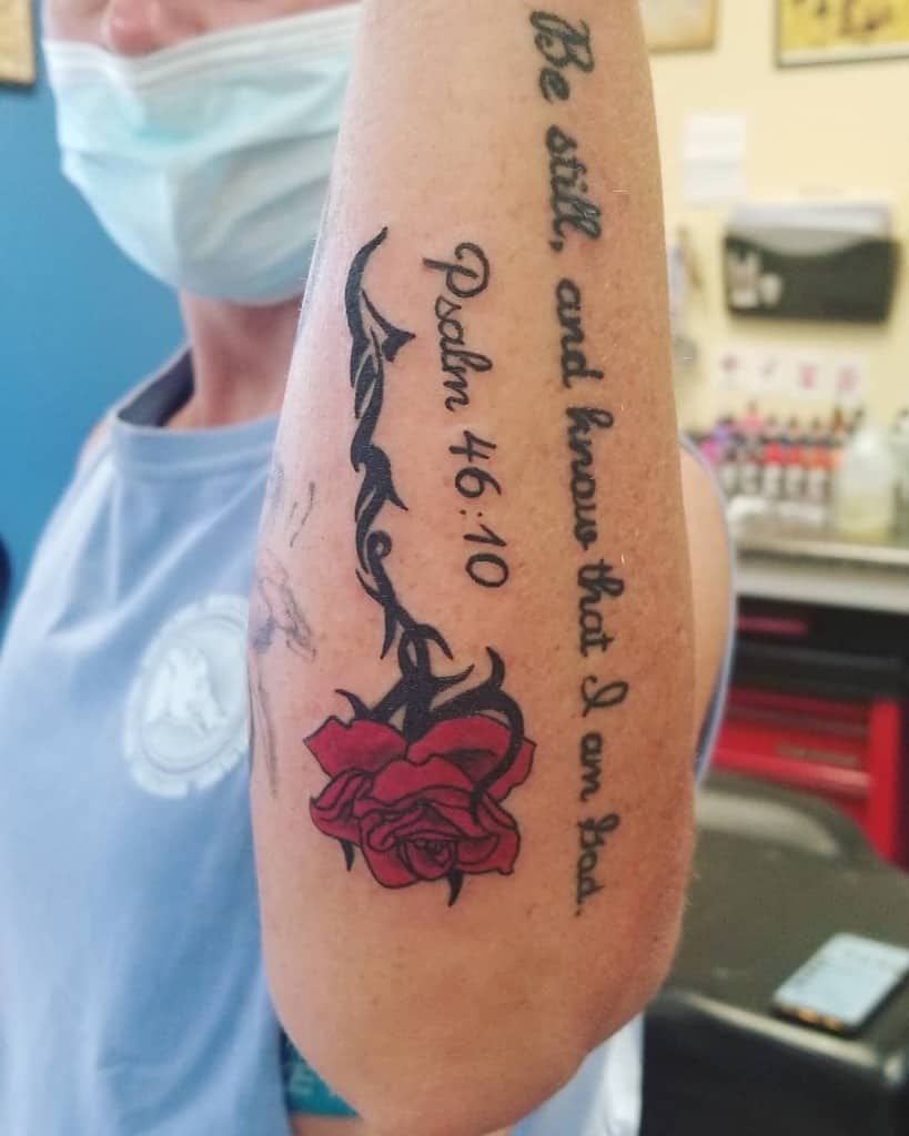 A person with a tattoo featuring Psalm 46:10, a red rose intertwined with a tribal vine on their arm