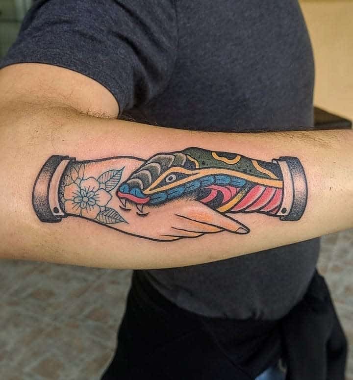 Colorful forearm tattoo of a handshake between a human and a snake, with a flower on the wrist of the human hand, in a bold traditional style
