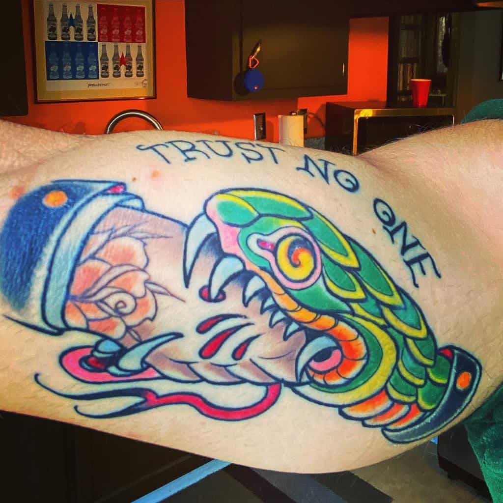 Vibrant forearm tattoo featuring a handshake between a human and a snake, with blood drops and the phrase 'Trust No One' above