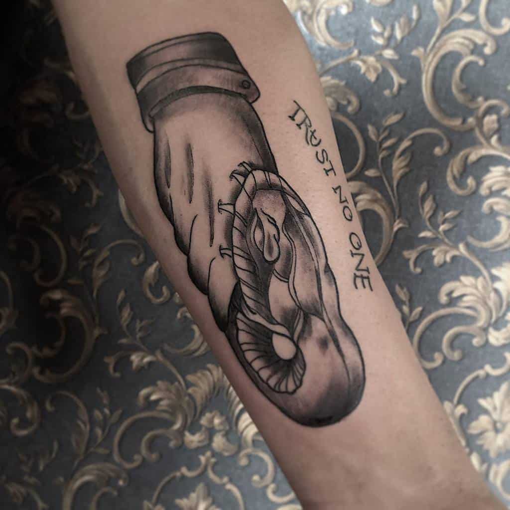 Black and gray forearm tattoo of a handshake between a human hand and a snake, with the phrase 'Trust No One' inked along the side