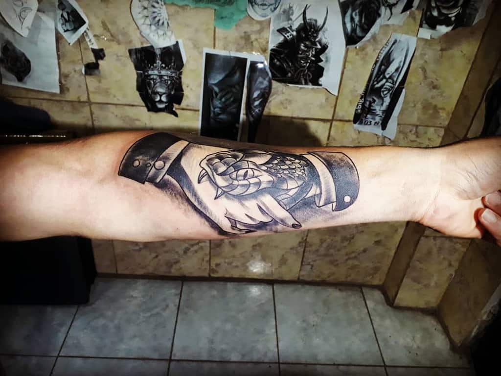 Detailed black and gray forearm tattoo of a handshake between a human and a snake, featuring bold shading and a traditional style