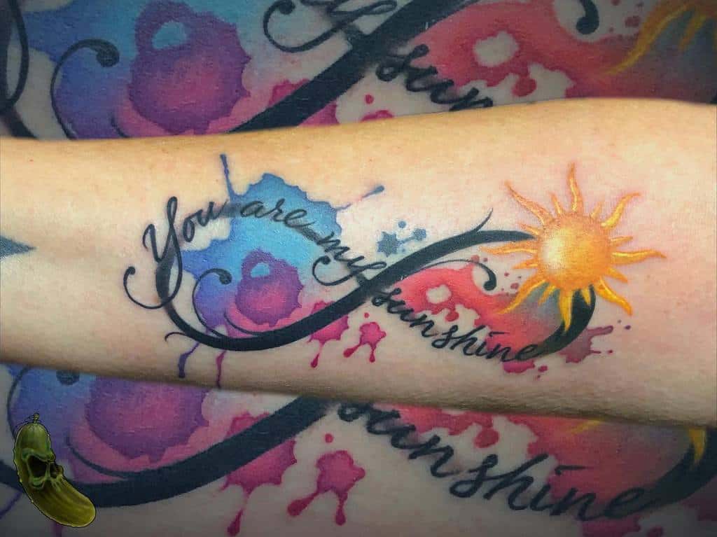 'You are my sunshine' tattoo with a vibrant sun and colorful watercolor splashes in pink, blue, and purple hues on the forearm