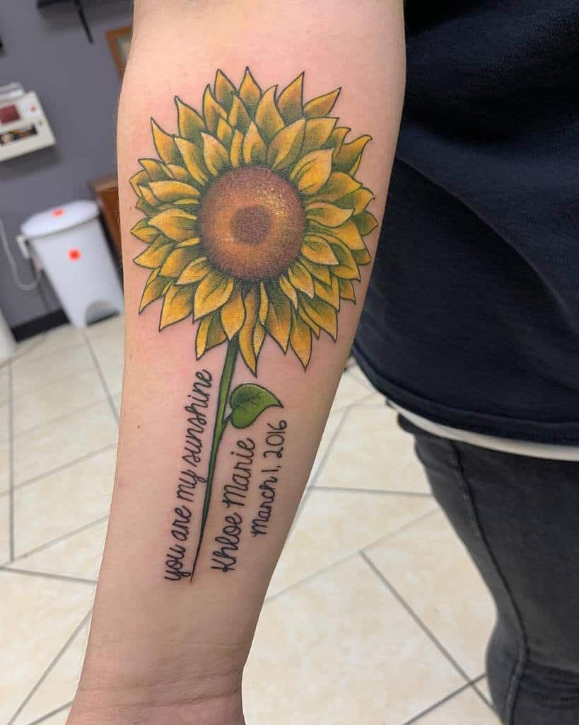 Tattoo of a sunflower with text on an arm, stating "You are my sunshine" and a name with date