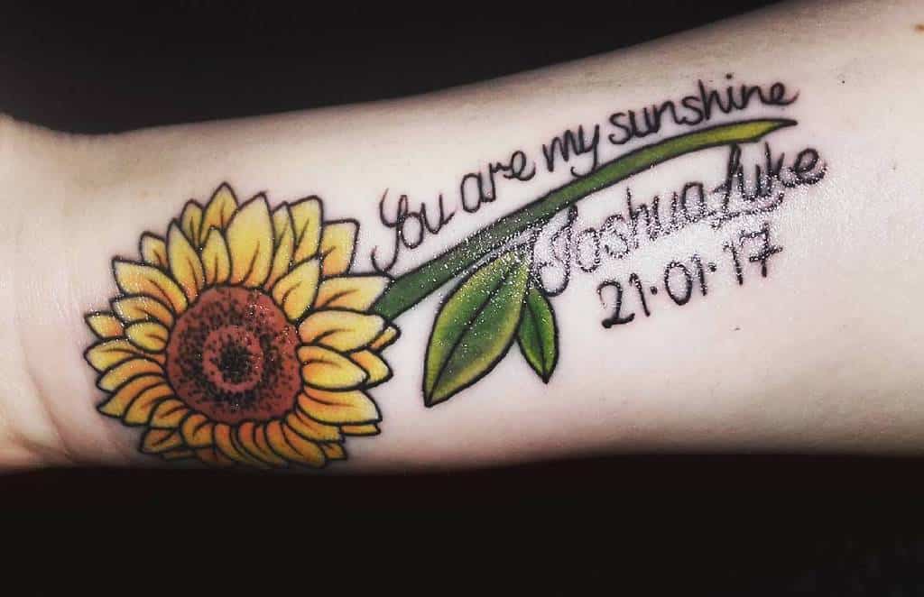 Forearm tattoo featuring a vibrant sunflower with 'You are my sunshine,' a name, and a date in elegant script