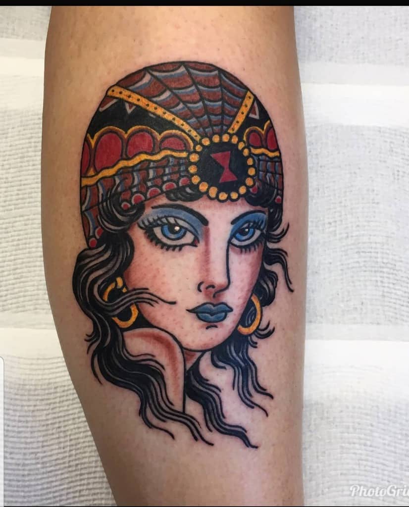Traditional tattoo of a gypsy woman with bold blue eyeshadow, a red and black ornate headpiece, and gold jewelry on the leg.