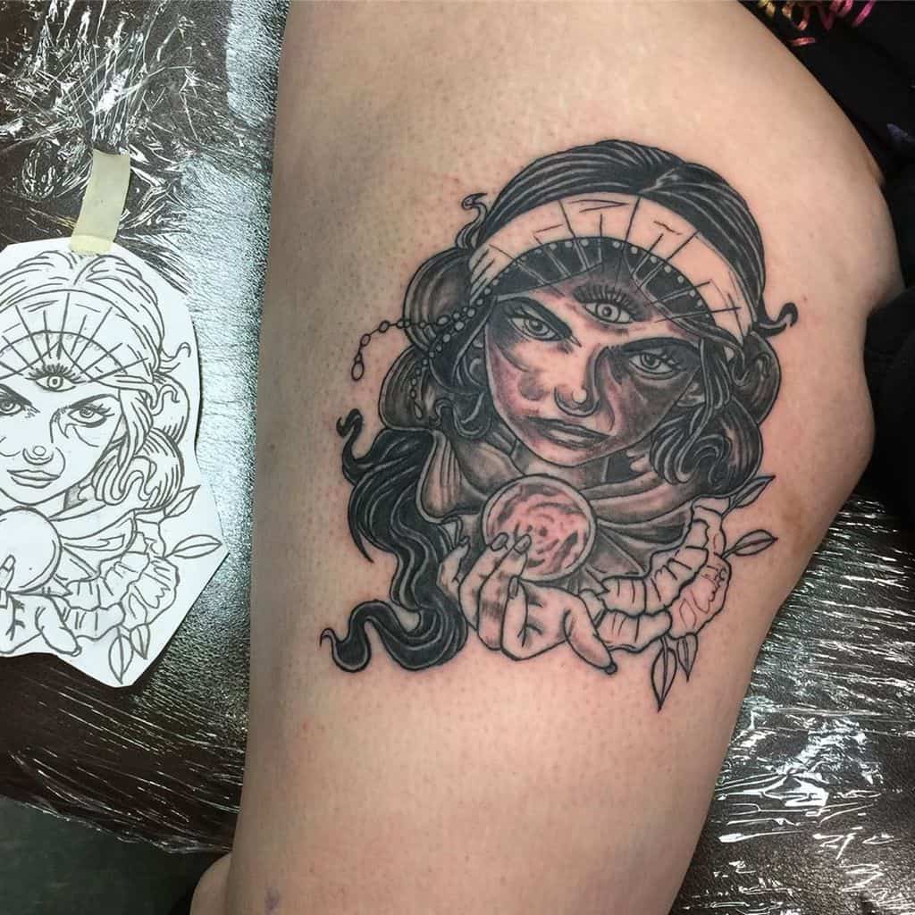 Black and grey tattoo of a mystical fortune teller with a third eye, intense shading, and a glowing crystal ball on the thigh.