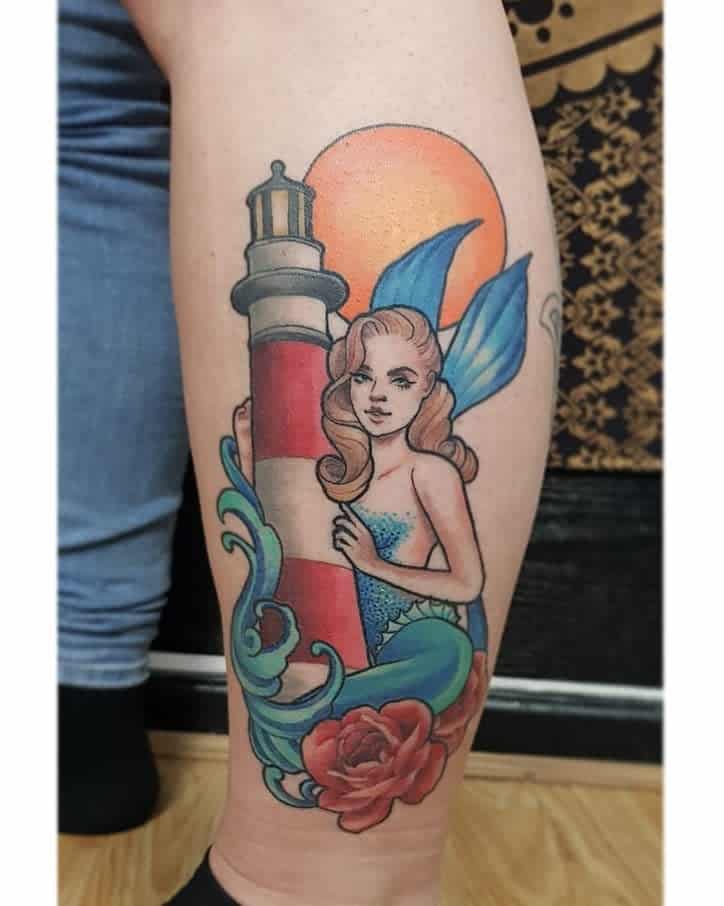 Colorful tattoo of a mermaid with blue fins leaning against a red and white lighthouse, with ocean waves, a rose, and a sunset.