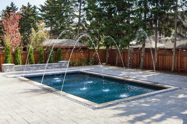 Modern backyard pool with arched water jets and paved surroundings.