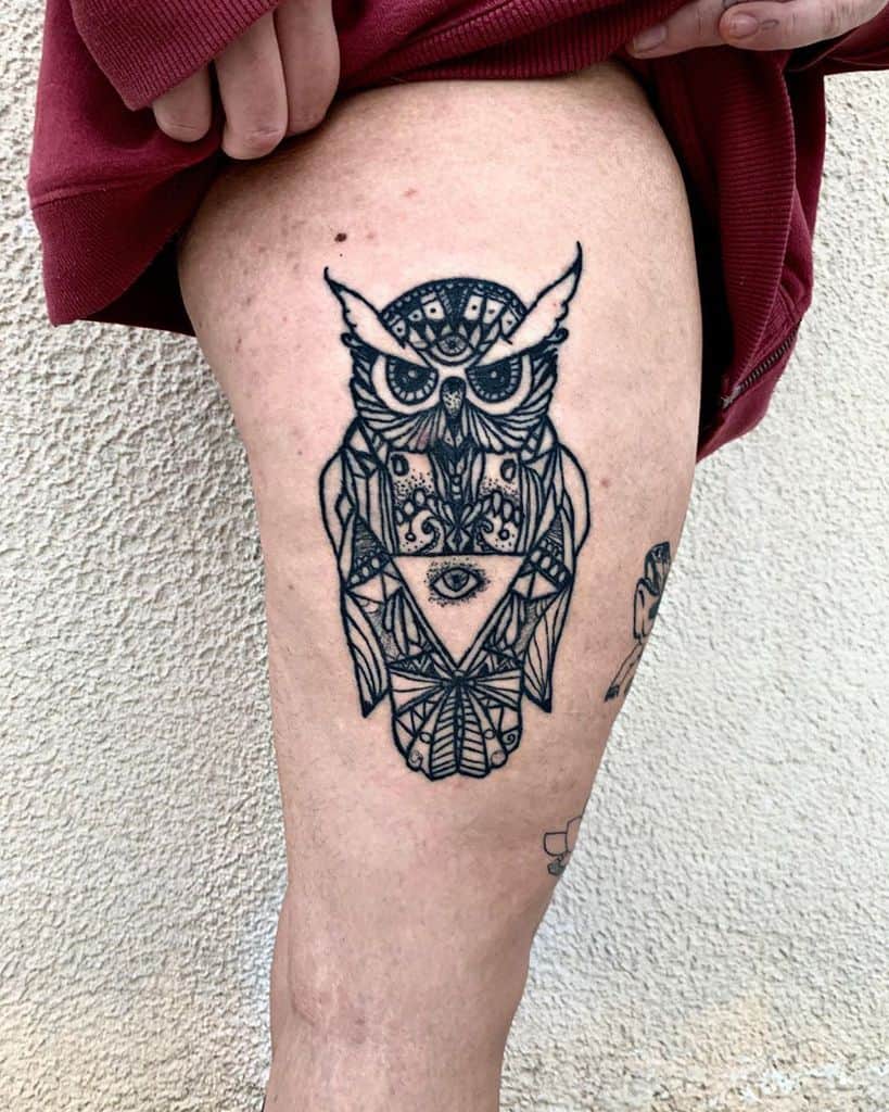 Tattoo of a geometrically detailed owl with an all-seeing eye on its chest, inked in black on the upper thigh.