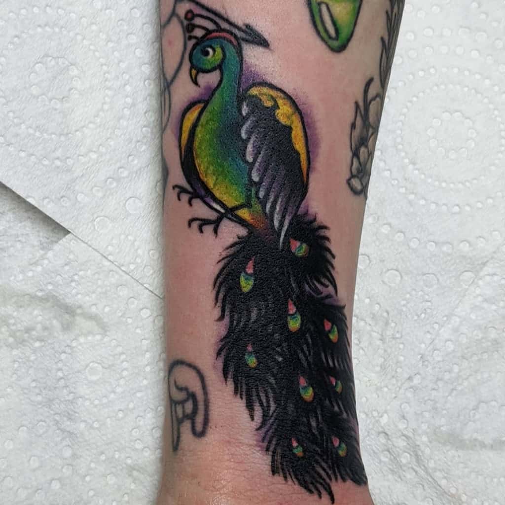 Colorful peacock tattoo on the forearm, featuring vibrant green and yellow tones with a dark, detailed tail adorned with small eye patterns.