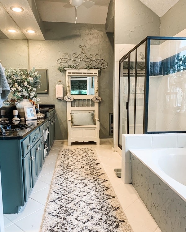 Elegant bathroom with teal cabinets, a black countertop, floral decor, large mirror, glass shower, and farmhouse charm