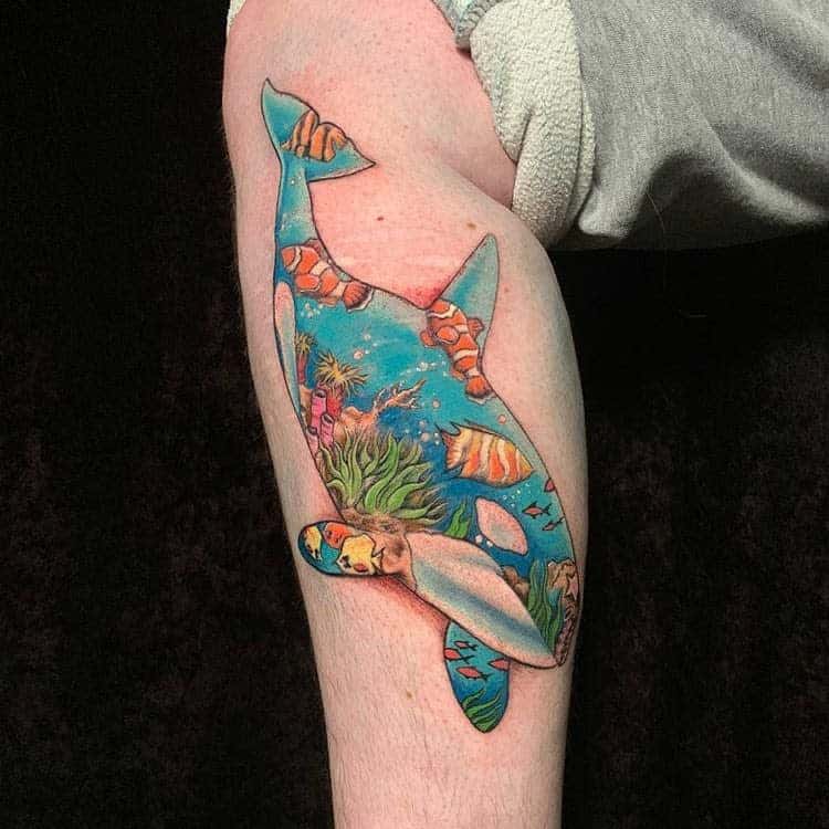 Colorful tattoo of an orca filled with ocean scene, featuring coral, fish, and sea plants, blending marine life within the whale’s silhouette