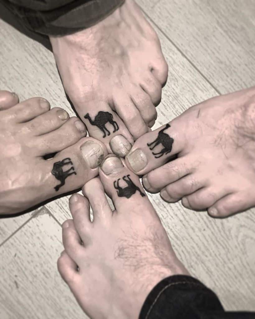 Four people with matching black camel tattoos on their big toes, arranged in a circle on a wooden floor for a fun group tattoo moment.