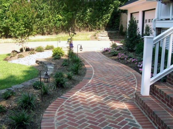 Curved brick walkway lined with plants and garden lights, leading towards a home with a white porch and lush greenery